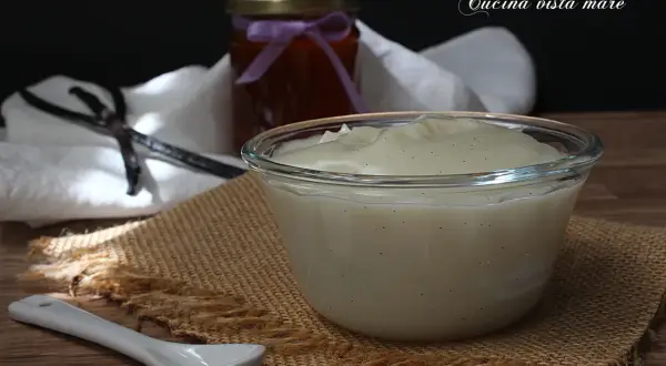 Una ciotola di cremosa crema al latte, profumata alla vaniglia, pronta per essere gustata. Accanto, un barattolo di marmellata e bacche di vaniglia.