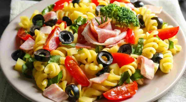 Un piatto di pasta fredda saporita con fusilli, pomodorini, olive nere, prosciutto cotto e un ciuffo di broccoli.