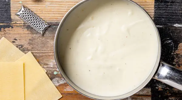 Besciamella ricetta: una cremosa salsa bianca in pentola, con sfoglie di pasta, noce moscata e grattugia su un tavolo di legno.