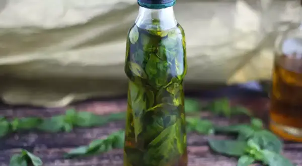 Bottiglia di vetro piena di olio al basilico, con foglie fresche sparse sul tavolo di legno.