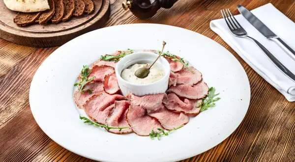 Fettine di arrosto rosa disposte a cerchio su un piatto bianco, con una ciotolina di salsa tonnata guarnita da un cappero.