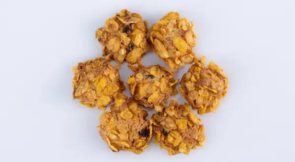 Biscotti croccanti con fiocchi di mais e uvetta, simili a rose del deserto, disposti a cerchio su sfondo bianco.