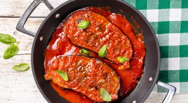 Melanzane in padella, succulente e cotte in un ricco sugo di pomodoro, guarnite con foglie di basilico fresco.