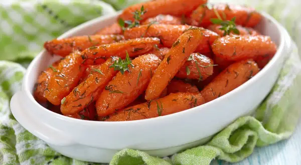 Carote glassate in padella, profumate con erbe aromatiche, servite in una ciotola bianca su un tovagliolo verde.