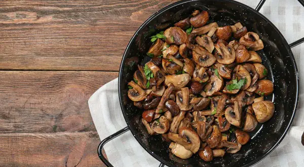 Funghi in padella, dorati e profumati, guarniti con prezzemolo fresco, pronti per essere gustati su un tavolo di legno rustico.