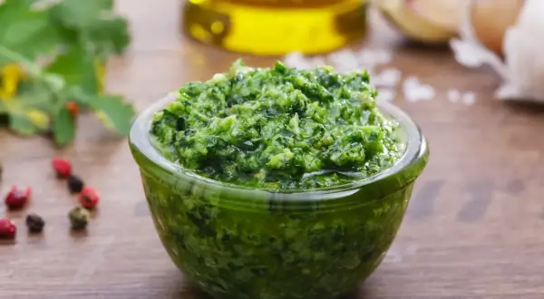 Una ciotola di fresca salsa verde, con ingredienti come aglio, pepe e olio d'oliva sullo sfondo.