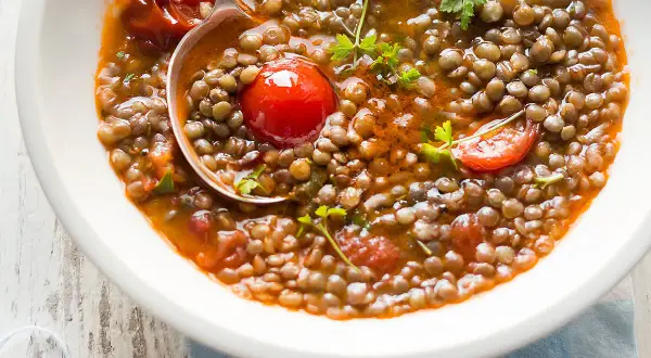 Una ciotola di zuppa di lenticchie fumante, con pomodorini e prezzemolo fresco. Un cucchiaio serve una porzione generosa di questo piatto confortante.