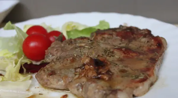 Una succulenta braciola di maiale, condita con erbe aromatiche, servita con pomodorini e insalata fresca su un piatto bianco.