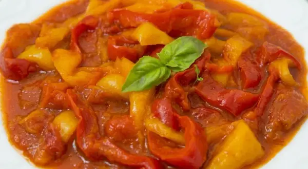 Peperonata ricetta della nonna: peperoni rossi e gialli stufati in un sugo saporito, guarniti con una fogliolina di basilico.