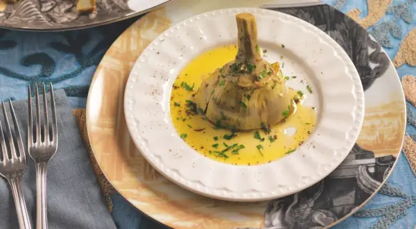 Un carciofo alla romana, tenero e profumato, servito su un piatto bianco con olio e prezzemolo.