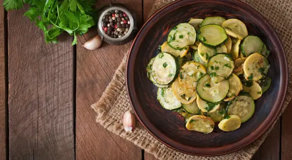 Piatto di zucchine trifolate, condite con aglio, prezzemolo e pepe, su un tavolo di legno rustico.