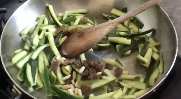 Zucchine tagliate a julienne con uvetta e pinoli in padella, pronte per essere trasformate in un delizioso piatto agrodolce.