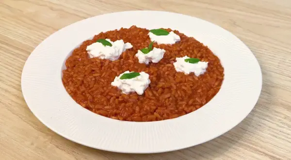 Un cremoso risotto al pomodoro, arricchito da ciuffi di ricotta fresca e basilico, servito su un piatto bianco.