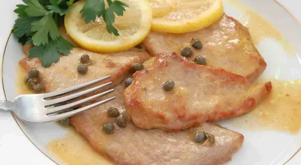 Scaloppine al limone cremose, guarnite con capperi, prezzemolo e fette di limone, pronte per essere gustate con una forchetta.