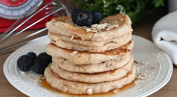 Pila di pancake con farina di avena, guarniti con mirtilli e sciroppo d'acero. Sullo sfondo, una bilancia rossa e un frullino.