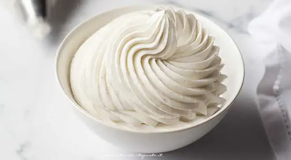 Una ciotola bianca piena di soffice crema chantilly, con striature perfette e puntini di vaniglia visibili.