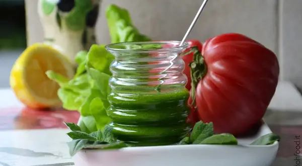 Un vasetto di salsa al basilico, con pomodoro, limone e lattuga sullo sfondo.