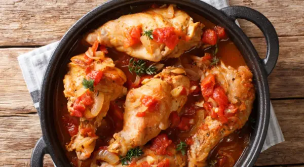 Coniglio all'ischitana in tegame, con pomodoro, cipolla e erbe aromatiche, servito su un tavolo di legno.