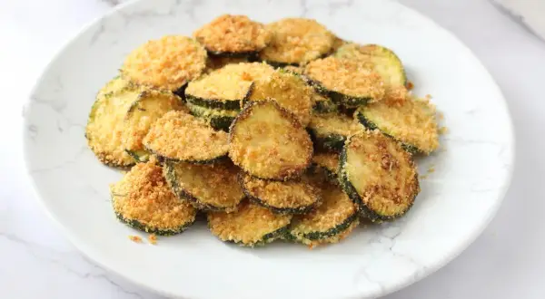 Fettine di zucchine impanate e cotte nella friggitrice ad aria, dorate e croccanti, servite su un piatto bianco.