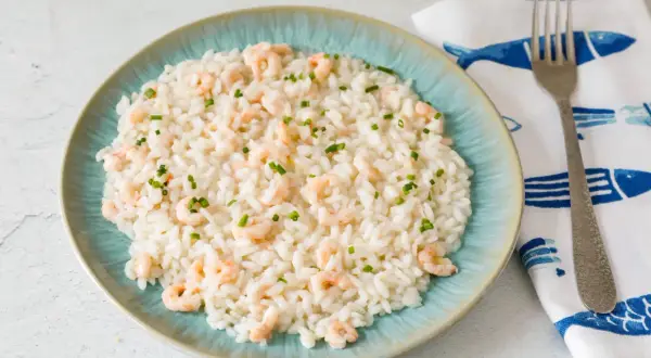 Un cremoso risotto gamberi e limone, guarnito con erba cipollina, servito su un piatto turchese. Accanto, un tovagliolo con pesci blu e una forchetta.