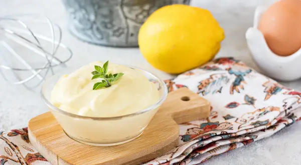 Una ciotola di cremosa maionese fatta in casa, guarnita con erbe fresche, accanto a limoni, un uovo e una frusta.