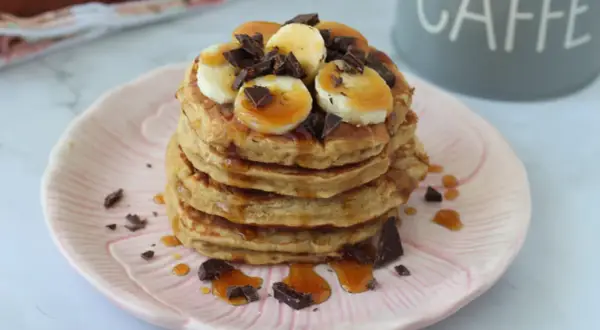 Pancake light con banana, cioccolato e sciroppo d'acero su un piatto rosa.