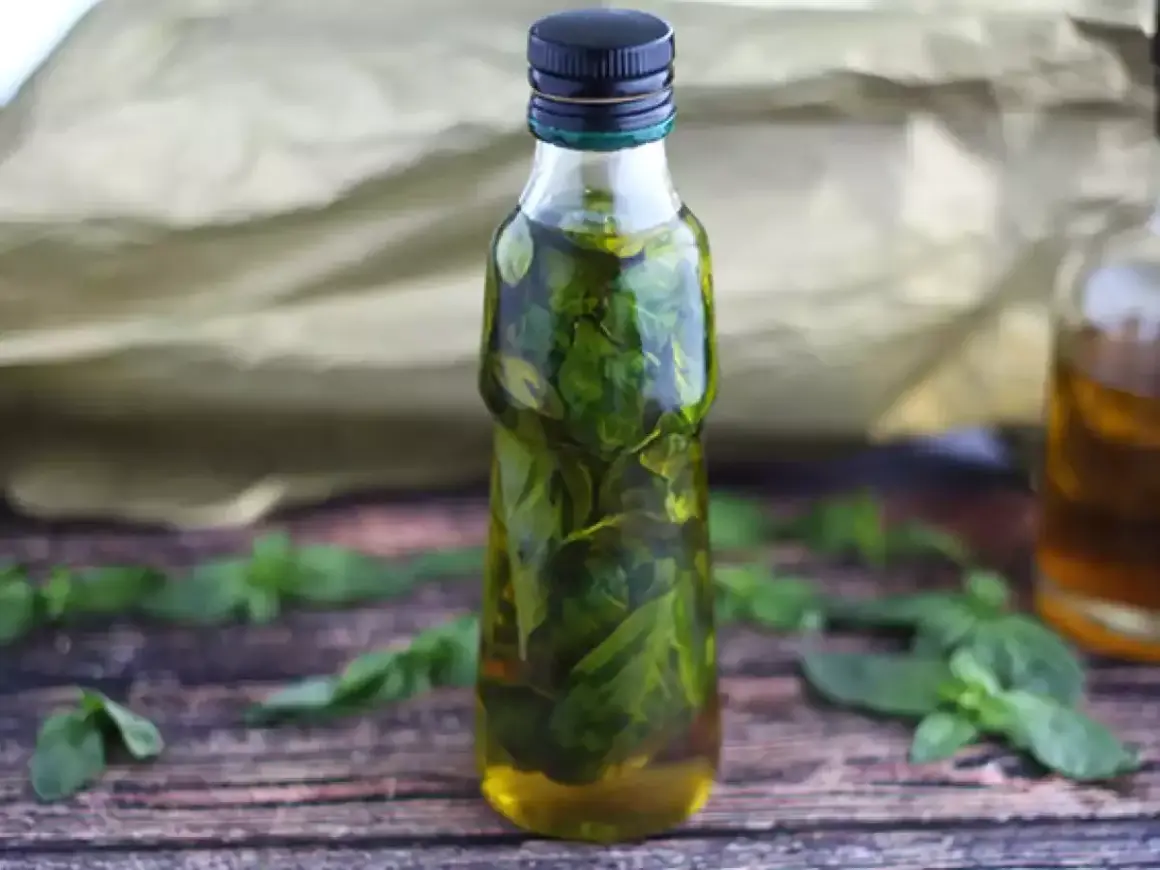 Bottiglia di vetro piena di olio al basilico, con foglie fresche sparse sul tavolo di legno.