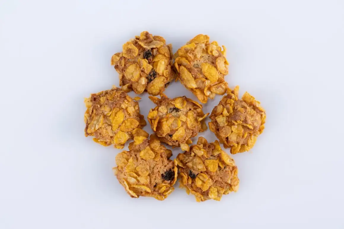 Biscotti croccanti con fiocchi di mais e uvetta, simili a rose del deserto, disposti a cerchio su sfondo bianco.