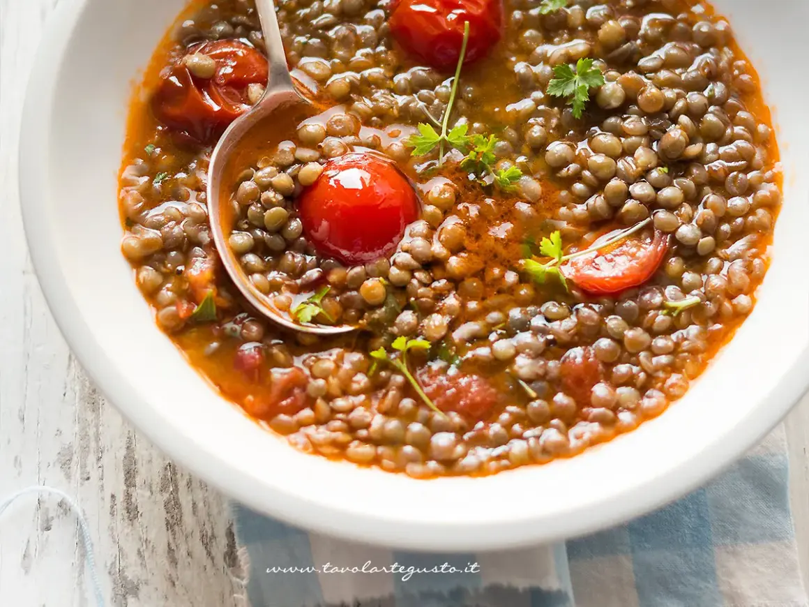 Una ciotola di zuppa di lenticchie fumante, con pomodorini e prezzemolo fresco. Un cucchiaio serve una porzione generosa di questo piatto confortante.