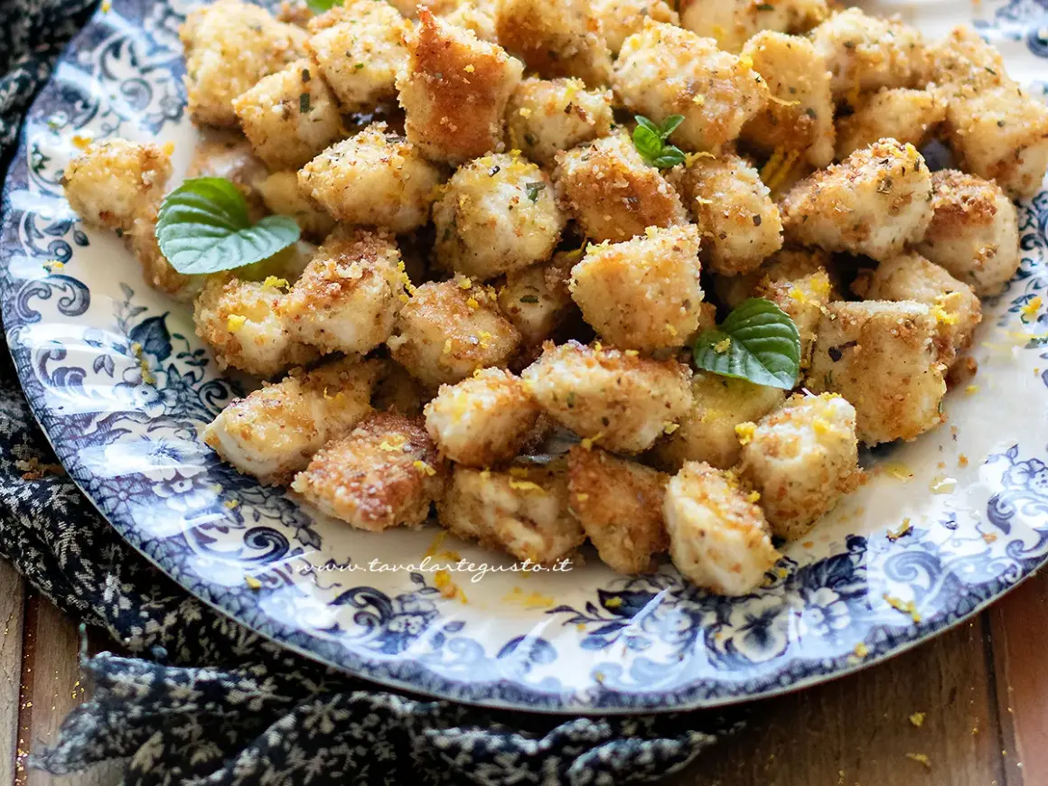 Bocconcini di pollo dorati e croccanti, guarniti con scorza di limone e foglioline di menta, serviti su un piatto decorato.