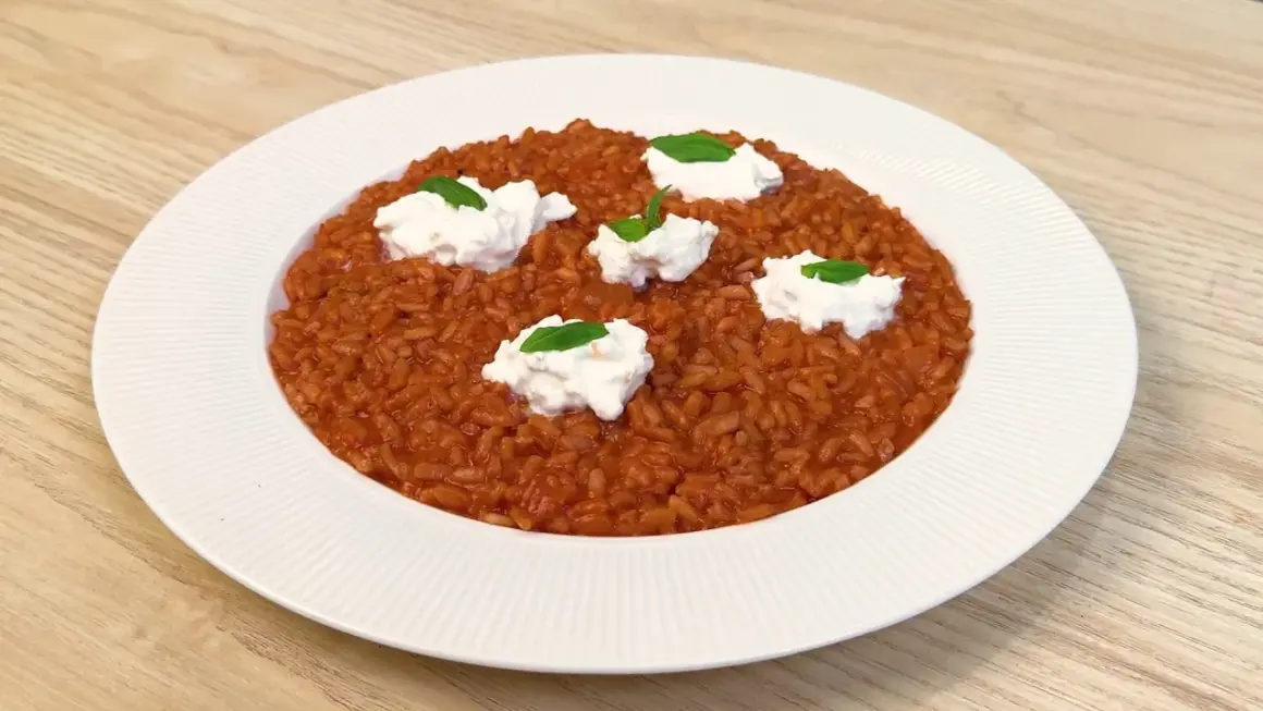 Un cremoso risotto al pomodoro, arricchito da ciuffi di ricotta fresca e basilico, servito su un piatto bianco.