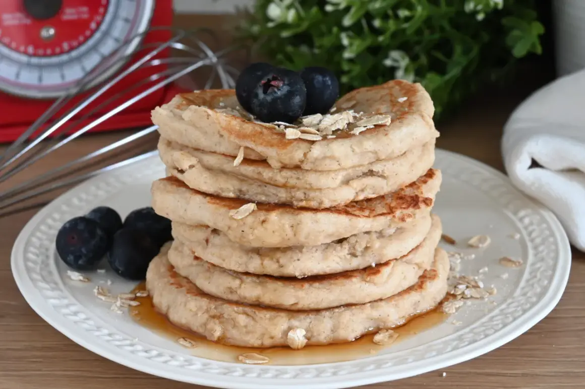 Pila di pancake con farina di avena, guarniti con mirtilli e sciroppo d'acero. Sullo sfondo, una bilancia rossa e un frullino.