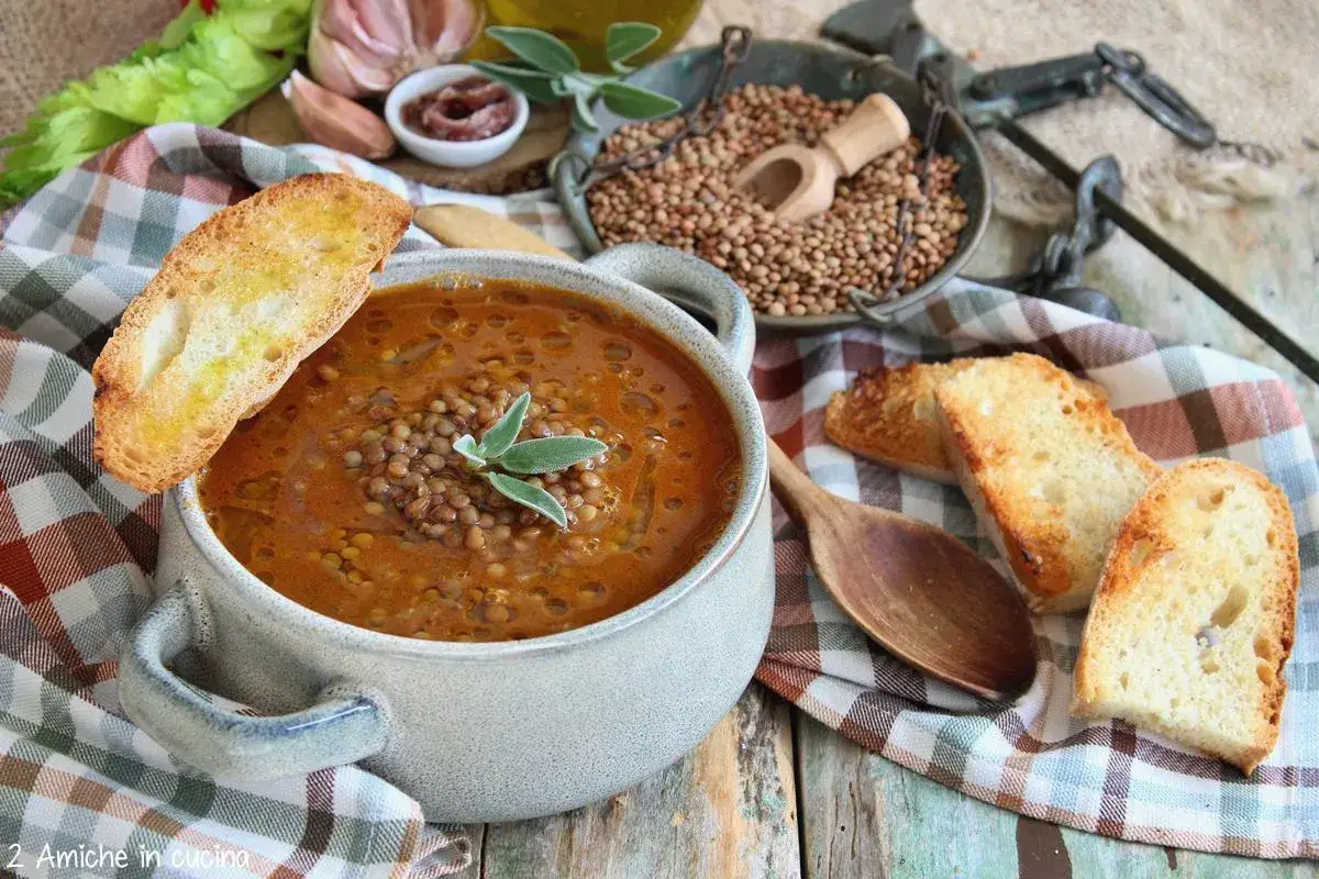 Una ciotola fumante di zuppa di lenticchie, guarnita con salvia, accompagnata da crostini dorati. Ingredienti freschi come aglio e sedano completano la scena.
