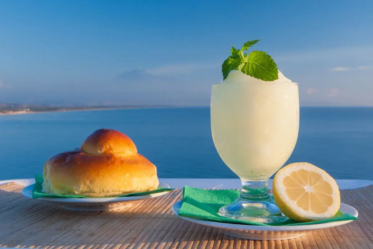 Una brioche e una rinfrescante granita al limone, guarnita con menta, su un tavolino con vista sul mare e sull'Etna.