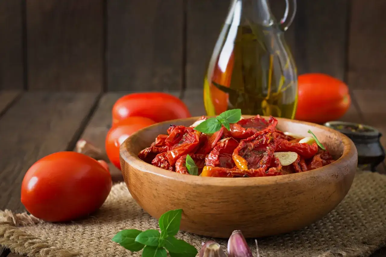 Pomodori secchi in una ciotola di legno, con pomodori freschi, aglio e olio d'oliva sullo sfondo.