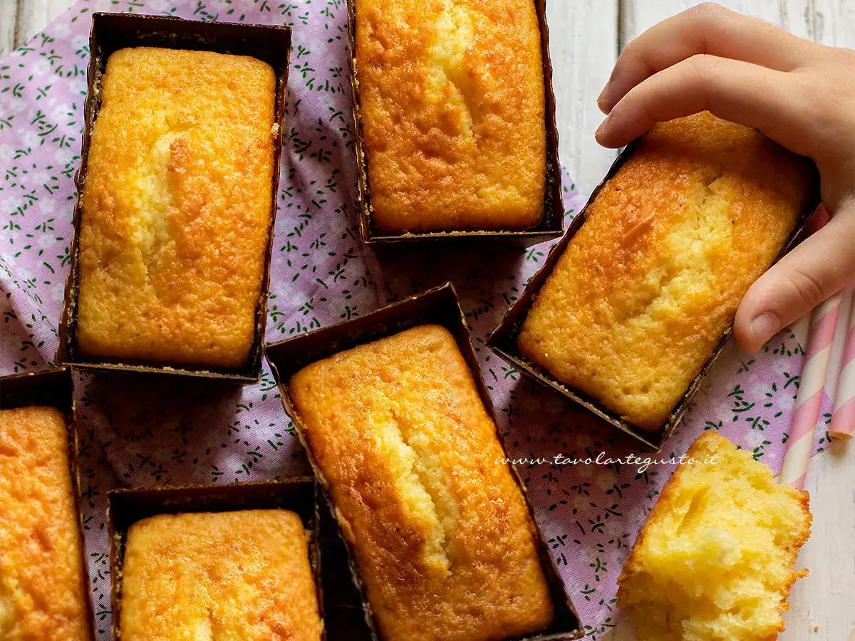 Plumcake yogurt dorati e soffici, pronti per essere gustati. Una mano ne prende uno, mentre un pezzetto mostra la morbidezza interna.