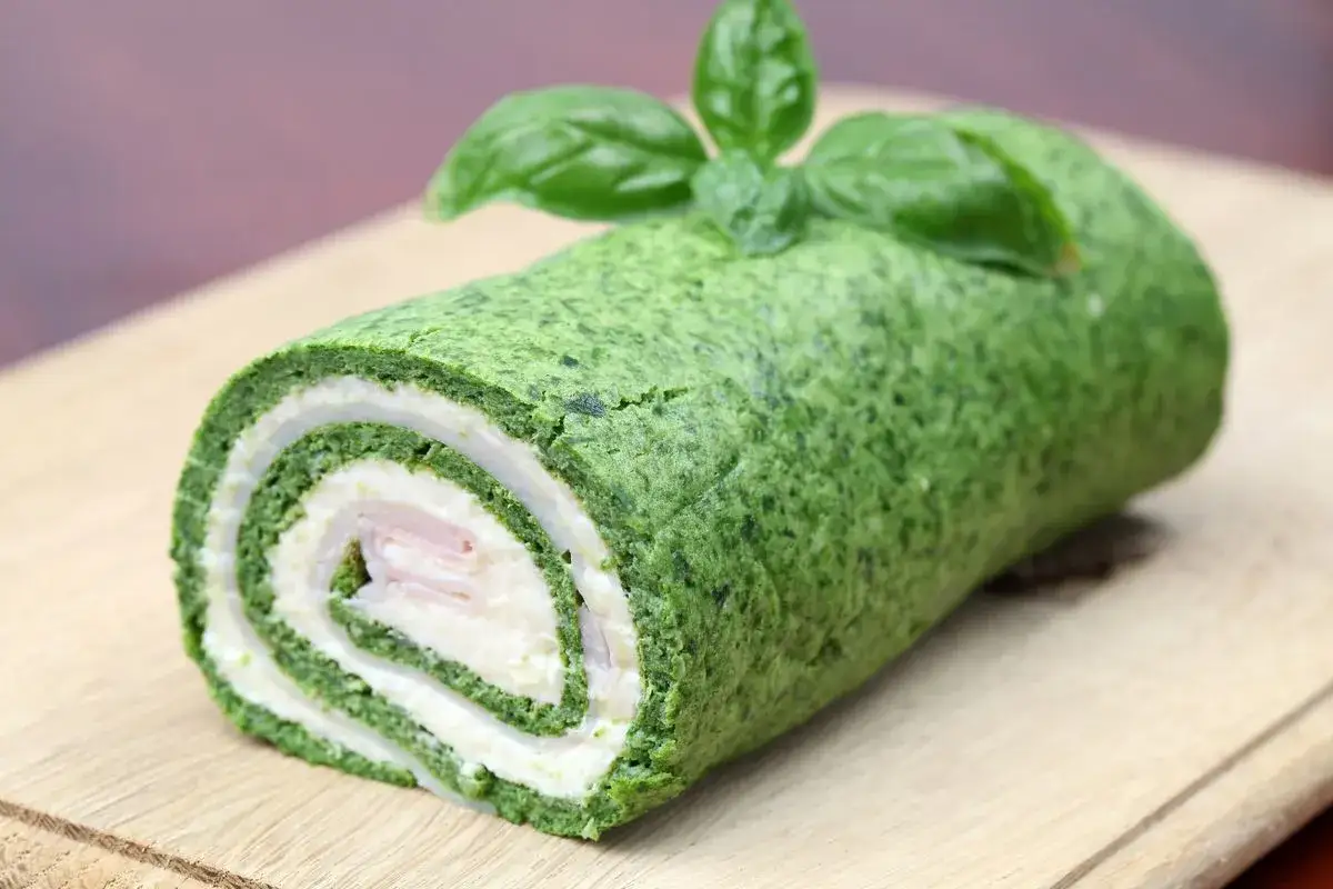 Rotolo di frittata verde con spinaci, ripieno di formaggio e prosciutto, guarnito con basilico fresco su tagliere di legno.