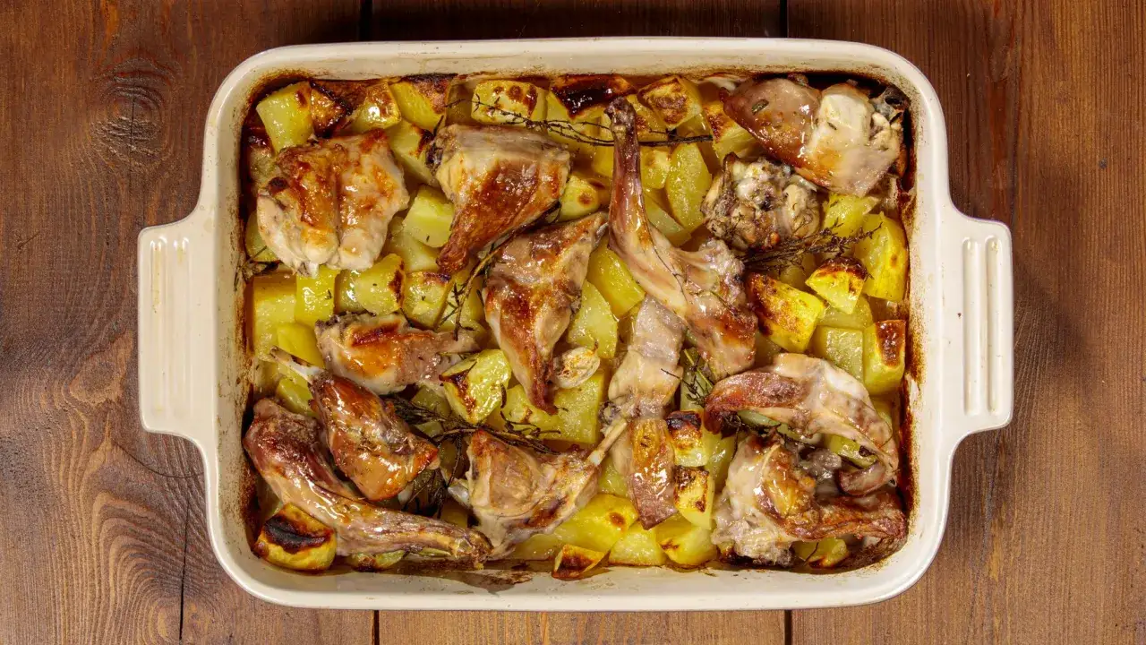 Delizioso coniglio al forno con patate dorate e rosmarino, servito in una teglia bianca su un tavolo di legno rustico.