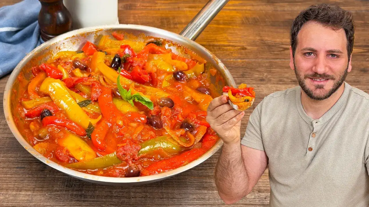 Un uomo sorride mostrando un crostino con peperonata ricetta della nonna, accanto a una padella piena di peperoni colorati e olive.