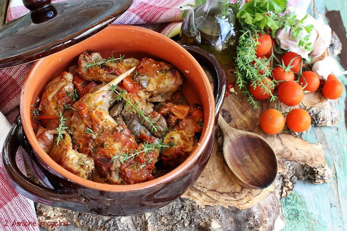 Un saporito coniglio all'ischitana in un tegame di terracotta, con pomodorini, erbe aromatiche e una bottiglia d'olio.