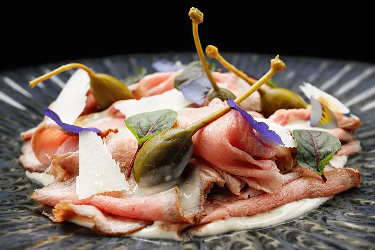 Fettine di arrosto con salsa tonnata, capperi, scaglie di parmigiano e fiori eduli su piatto scuro.