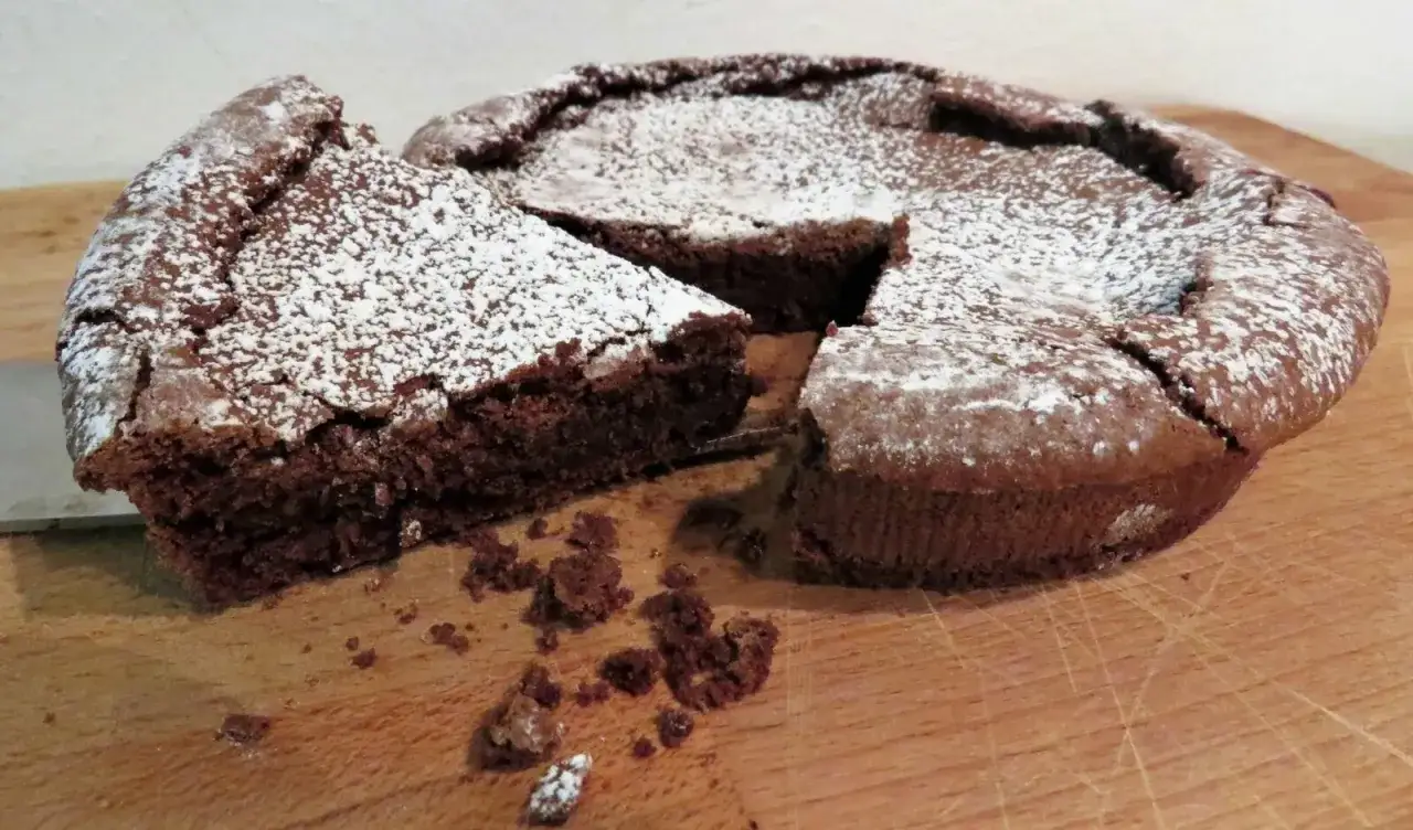 Una fetta di torta tenerina al cioccolato, cosparsa di zucchero a velo, su un tagliere di legno.