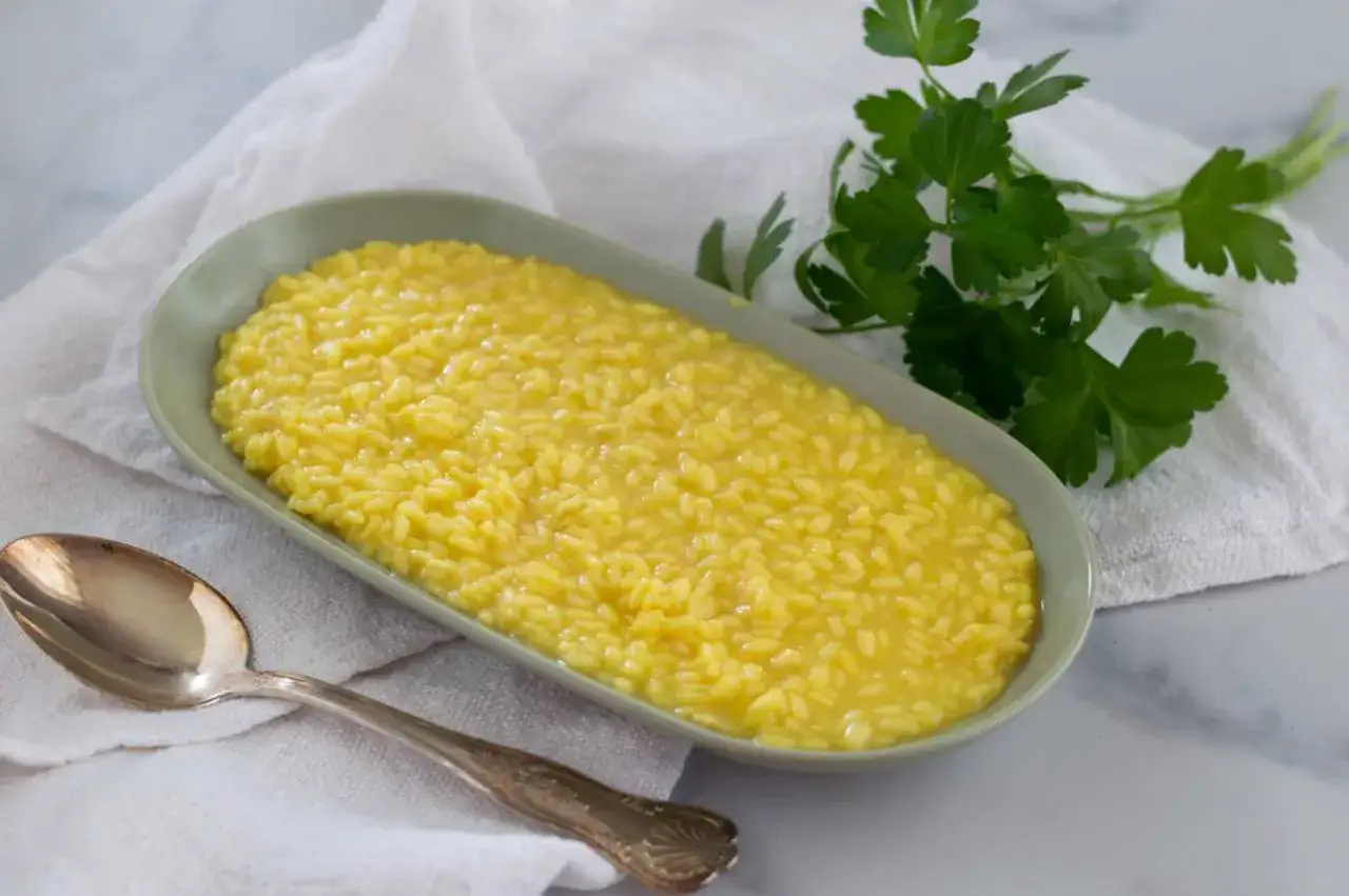 Un cremoso risotto alla milanese, giallo intenso, servito in un piatto ovale con un cucchiaio d'argento e un rametto di prezzemolo fresco.