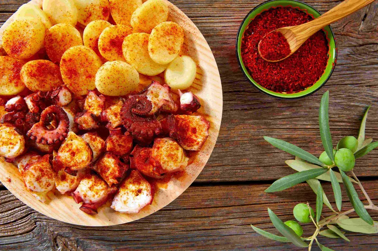 Piatto di polpo alla gallega con patate e paprika, accompagnato da olive verdi su un tavolo di legno.