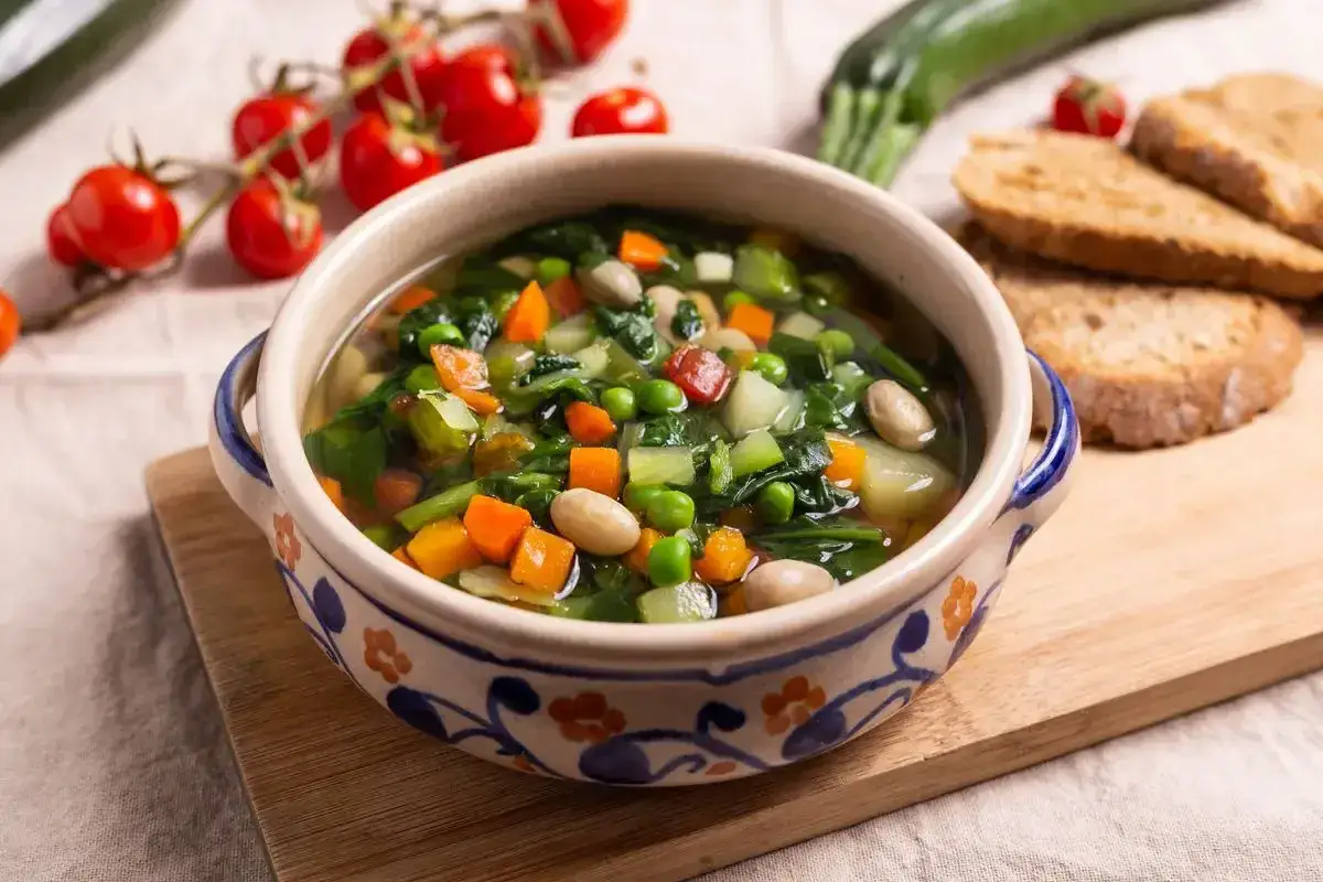Un fumante minestrone di verdure in una ciotola decorata, con pomodorini, zucchine e pane tostato sullo sfondo.