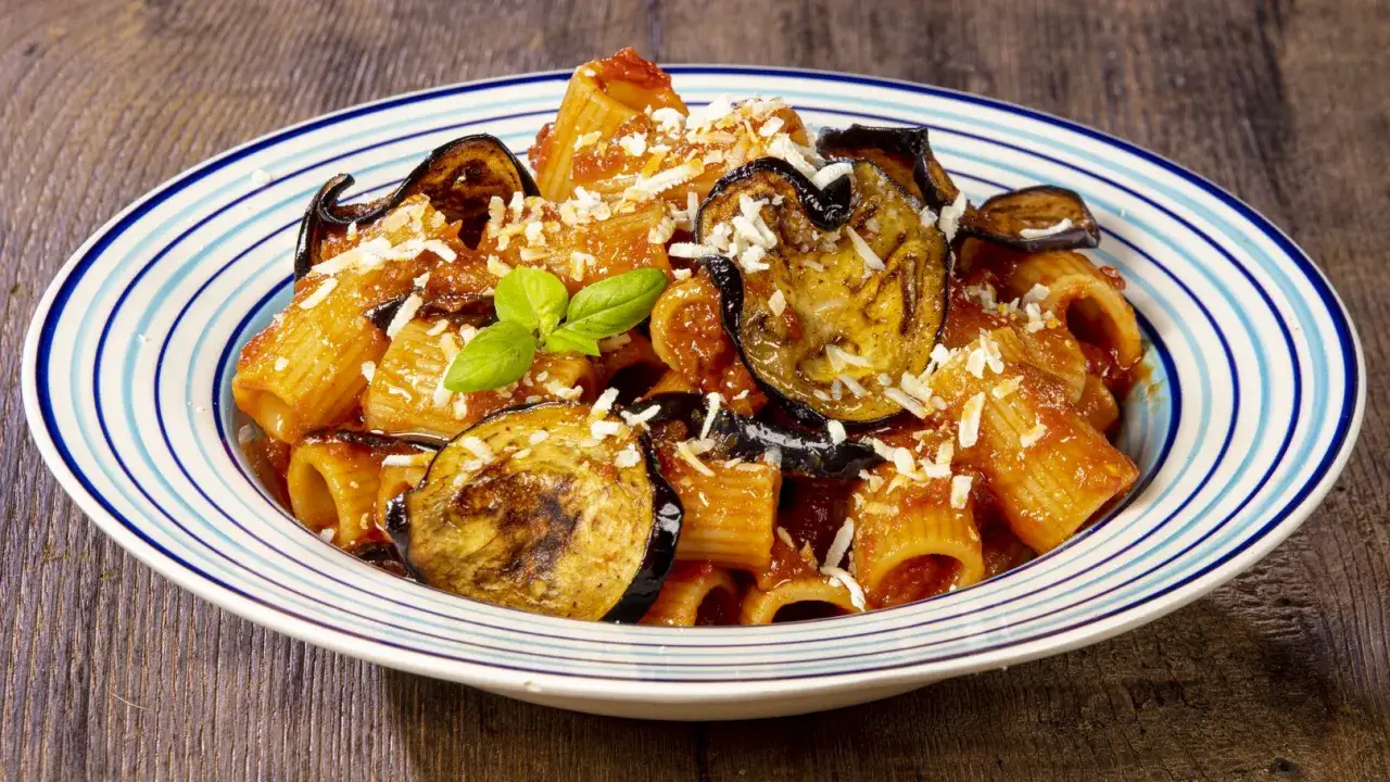 Un piatto di pasta alla norma con melanzane fritte, sugo di pomodoro e ricotta salata grattugiata, guarnito con basilico fresco.