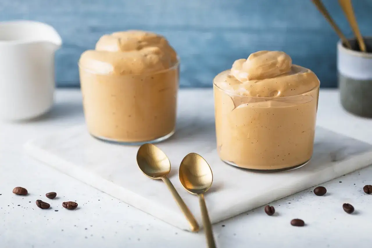 Due bicchieri di cremosa schiuma di caff&egrave;, perfetti per una ricetta crema di caff&egrave; senza panna. Due cucchiaini dorati e chicchi di caff&egrave; completano la scena.
