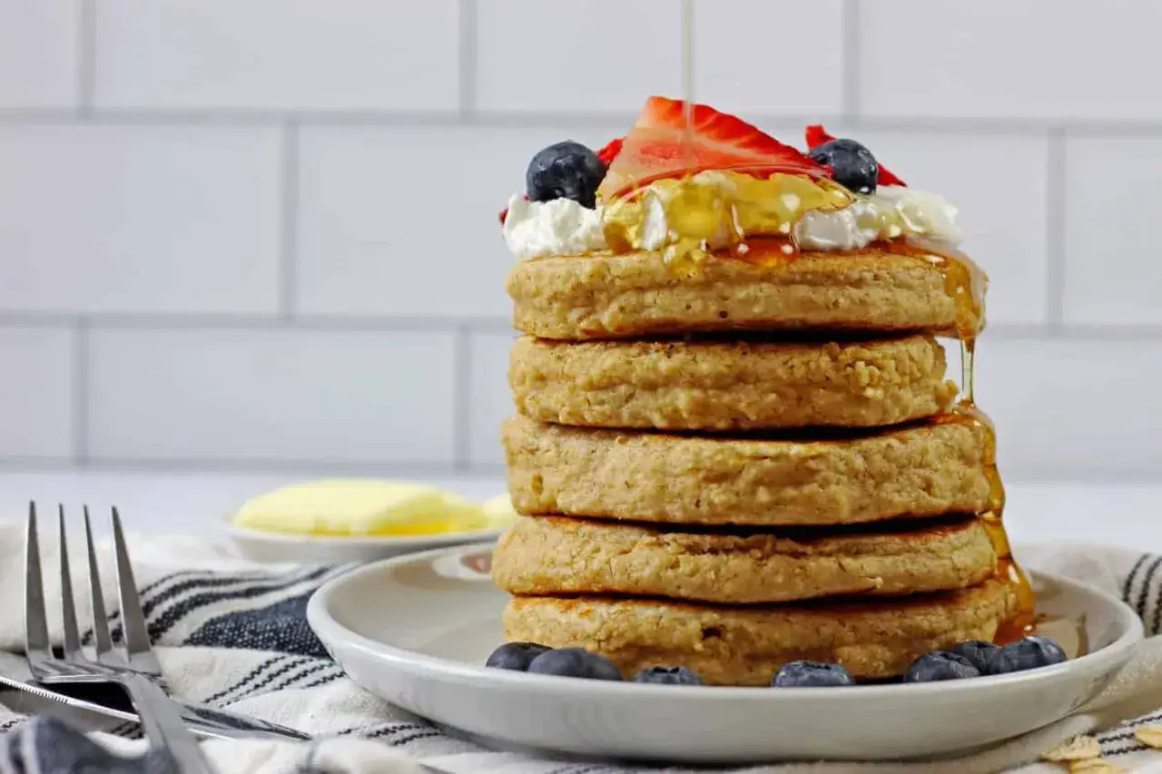 Pila di pancake con farina di avena, guarniti con panna, fragole, mirtilli e sciroppo d'acero che cola.