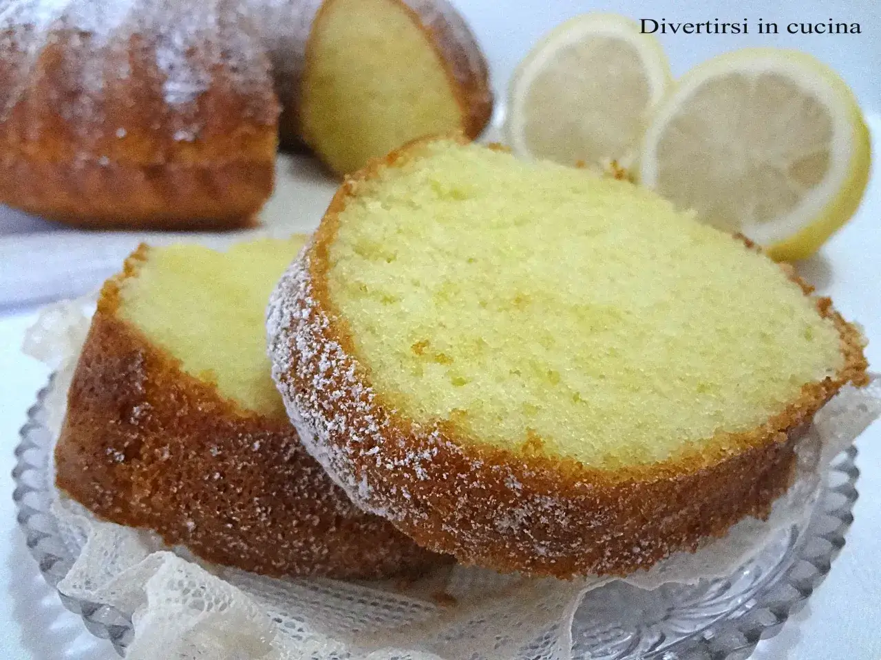 Fette di soffice plumcake al limone, spolverate di zucchero a velo, con spicchi di limone sullo sfondo.