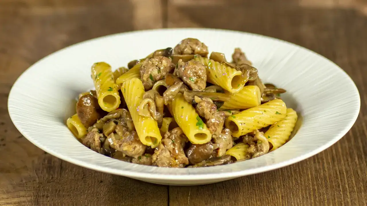 Piatto di pasta salsiccia e funghi, con rigatoni conditi con pezzi di salsiccia e funghi trifolati, guarnito con prezzemolo tritato.